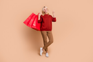 Full length photo of sweet friendly woman wear red sweater showing v-sign rising shoppers isolated beige color background