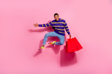 Full length photo of cheerful good mood man wear striped sweater jumping high holding shoppers isolated pink color background