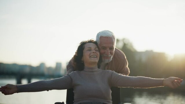 Elderly husband walking with wife in wheelchair. Senior family couple spending time in city park. Man caring woman showing love, they talking happy together. Living with physical disability concept.