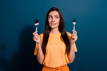 Portrait of biting lips woman wear holding metal spoon and fork looking empty space choose dish isolated on dark blue color background