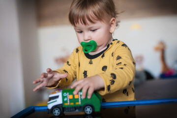 Cute baby boy with pacifier in mouth plays with a small toy truck, driving it around a make-believe...