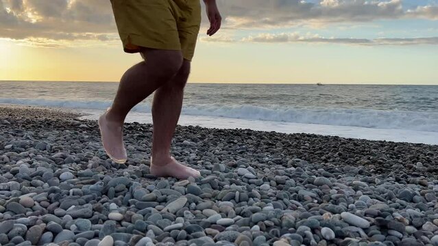 4K video. White adult man with hairy bare legs in yellow shorts, protective transparent shoes walks along pebble beach in evening against backdrop of sunset, setting sun, sea waves. Travel, tourism