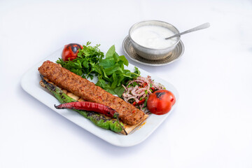 Top view of traditional grilled Adana kebab with cooked spicy shish kebab, pepper, tomato, and fresh greenery on a white plate with a metal pot of foamy ayran isolated on white background.