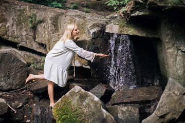 A smiling happy pregnant girl in a light vintage dress, with blond short hair, stands against the backdrop of picturesque rocks near a waterfall in the forest.