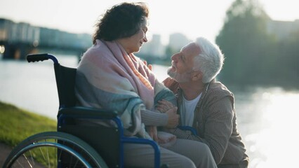 Husband walking with wife in wheelchair. Senior family couple spending time in city park near pond. Man hugging female showing love care help, they smiling. Living with physical disability concept.