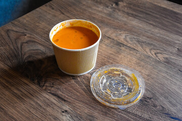Tomato soup in a paper package on a wooden table. Paper bag. Takeaway food. Plastic cap. Lunch delivery