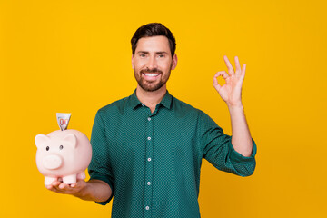 Photo of optimistic pleasant man dressed dotted shirt put dollar banknote in money piggy box show...