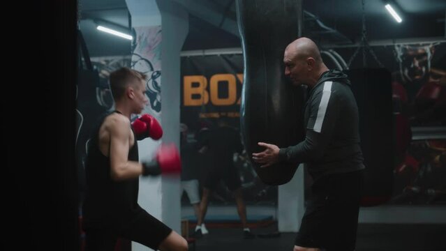 Young boxer in boxing gloves exercises with trainer in dark gym before fighting competition. Athletic teen hits punching bag while practicing in boxing gym. Physical activity and intensive workout.