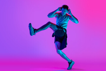 Fototapeta na wymiar Young muscular man in sportswear, wearing headphones and training against gradient pink background in neon light. Concept of active and healthy lifestyle, sport, fitness, endurance