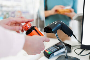 Close up shot of female pharmacist's hands holding medicament or drug and using barcode reader on cash register. Unrecognizable customer in a background.