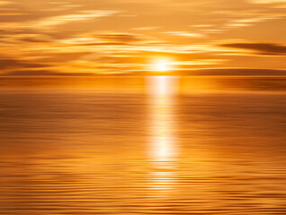 A motion blurred sunrise or sunset with the sun on the horizon casting a white ribbon of light over golden water. Bright yellow, orange, gold, and slivers of warm clouds fill the frame. 