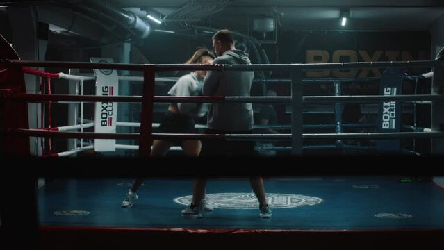 Female fighter in boxing gloves practices fighting technique with male trainer and exercises before championship in dark gym. Boxer hits punching mitts on boxing ring. Physical activity and workout.
