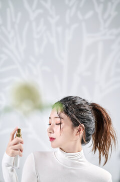 A young, beautiful Asian girl closes her eyes in front of a wall with a shadow of a tree and sprays her face with spray-type cosmetics