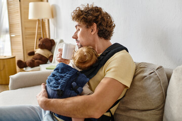 curly haired man with infant son in baby carrier holding cup of tea in living room, fatherhood