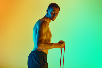 Athletic young man, with muscular, relief shirtless body training with resistance band against gradient blue yellow background in neon light. Concept of active and healthy lifestyle, sport, fitness