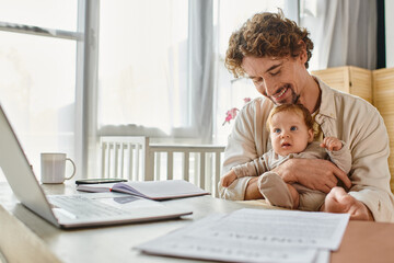 happy father holding infant son while working from home near papers and gadgets, work-life balance