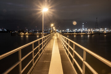 This image captures the quiet allure of a well-lit pier stretching into the distance, with the night-time activity of a city port in the background. The bright lamplight guides the eye along the