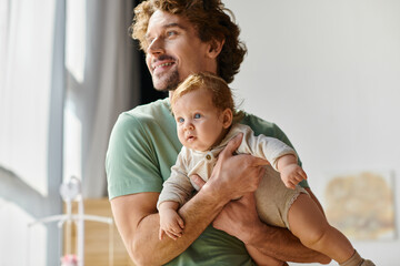 curly-haired and pleased father holding in arms his infant son in cozy bedroom at home, fatherhood