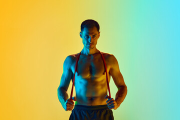 Handsome young man with muscular, shirtless, relief body posing wit fitness resistance band against gradient blue yellow background in neon light. Concept of active and healthy lifestyle, sport