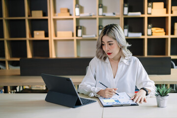 Confident beautiful Asian businesswoman typing laptop computer and digital tablet while holding coffee at office.
