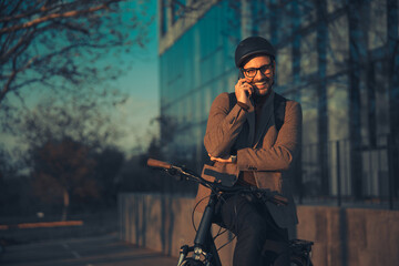 A cheerful young businessman casually sitting on a bicycle while having a phone call outside