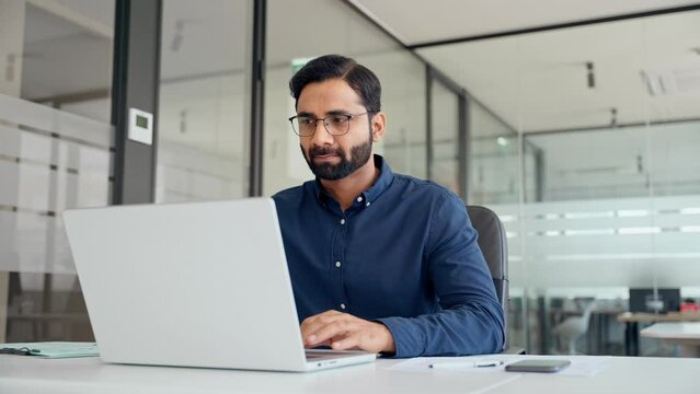 Busy Indian professional business man employee using laptop working in office. Professional financial analyst typing on computer technology working with ai solutions sitting at office desk. - Powered by Adobe