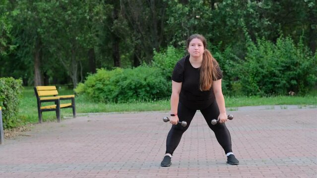 Overweight girl doing sports outdoors, lifting dumbbells.