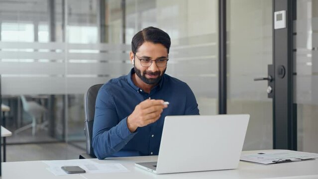 Happy Indian business man hr manager wearing earbud waving hand having hybrid virtual interview meeting, talking to client consulting customer on conference call working on laptop computer in office.