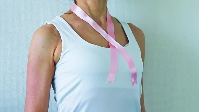 Shot of the woman in the white top, with pink ribbon on her neck as a symbol of breast cancer awareness, performing self examination of the breasts, looking for abnormalities. Healthcare