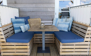 This image presents a cozy corner booth seating setup, characteristic of a beach house patio or a seaside cafe. The natural wood table and bench frames, combined with plush blue striped cushions