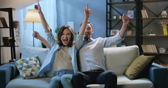 Excited couple sport fans watch the game on TV at home together, celebrating victory, screaming, raising hands up, hugging. Man and woman celebrating a win