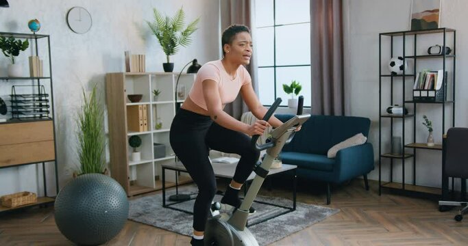 Attractive smiling athletic fit african american in sportswear woman exercising on stationary bike during morning home workout