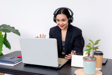 Cheerful female office employee using a laptop while working at her desk with headphones, coffee,...