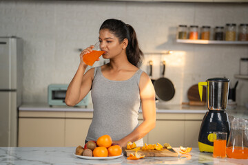 Young Indian woman in active wear drinking orange juice by drinking at kitchen - concept of healthy...