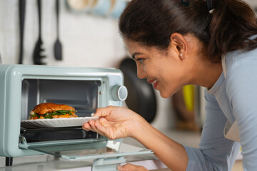 Happy Indian woman cooking burger by placing it on microwave oven at kitchen - concept of healthy eating, domestic lifestyle and technology