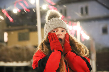 An 8-year-old girl on the background of lights in red gloves holds her hands near her face wondering at the snow