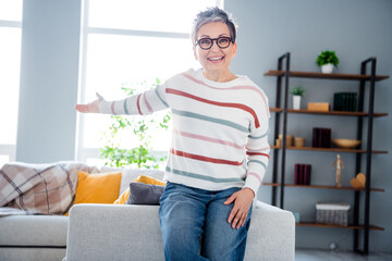 Photo of pretty excited woman dressed striped sweater eyewear sitting couch showing arm apartment indoors house home room