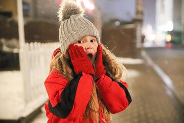 Adorable little girl surprises herself against the background of snow in winter in gray hat and dirty jacket