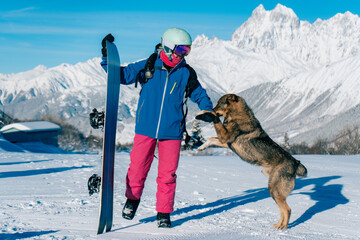 Female snowboarder playing with dog on ski resort, winter sport outdoor, sunny day in mountains