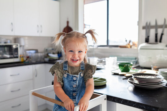 Happy toddler girl standing in messy kitchen with dishes piled on bench