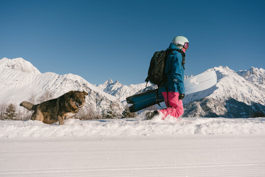 Female snowboarder freerider  hiking with dog on ski resort, winter sport outdoor, sunny day in mountains