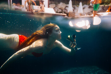 A beautiful red-haired woman poses with a cocktail under water.