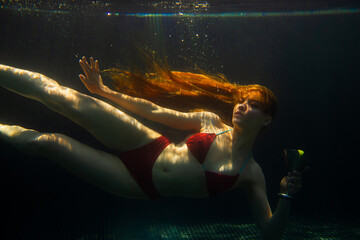 A beautiful red-haired woman poses with a cocktail under water.