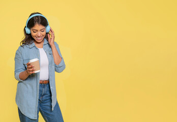 a happy dancing woman with blue  headphones  in jeans and denim shirt drinking coffe on yellow...