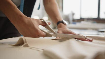 Young male tailor cutting fabric with scissors at table in dressmaking workshop. Tilt-up shot - Powered by Adobe