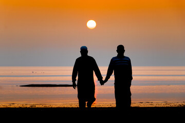 Silhouette view of two Muslim men walking hand in hand on a sea beach during twilight sunset. Friendship is an immortal concept even in old age. - obrazy, fototapety, plakaty