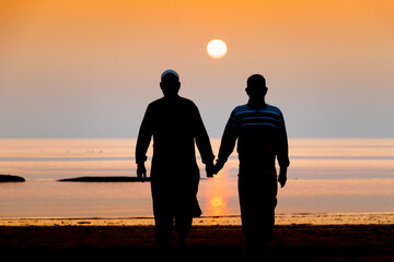 Silhouette view of two Muslim men walking hand in hand on a sea beach during twilight sunset....