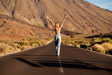 Fototapeta na wymiar Back view of young woman walking along empty asphalt road in the middle of volcanic area with raised hands, topless. Freedom, travel concept.