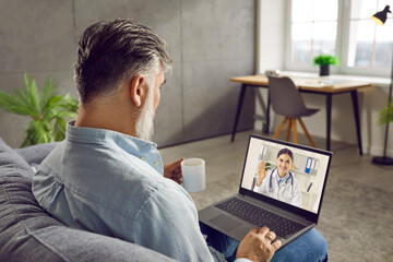 Senior man having online medical consultation via video call. Mature man sitting on sofa at home, drinking coffee, using laptop computer and having online conference with remote doctor