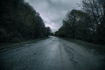Old gloomy road near trees. Bad asphalt. Gloomy atmosphere. Evening.